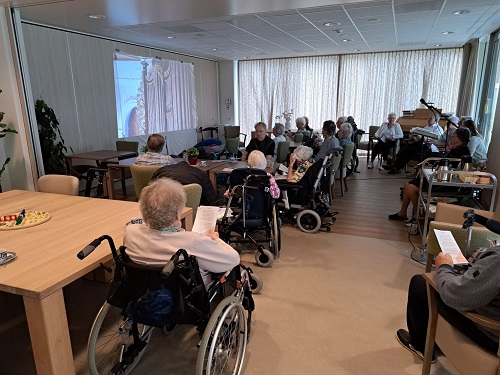Een groep ouderen in rolstoelen kijkt naar een presentatie in een gemeenschappelijke ruimte met een piano en tafels.
