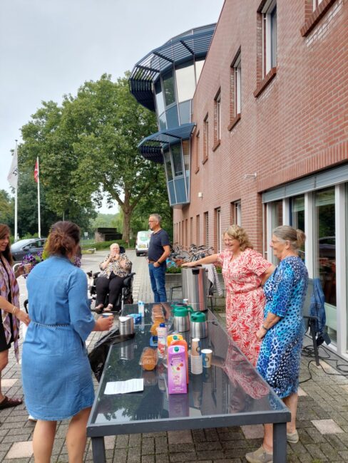 Groep mensen verzameld rond een tafel buiten een bakstenen gebouw, met koffie en snacks, op een bewolkte dag.