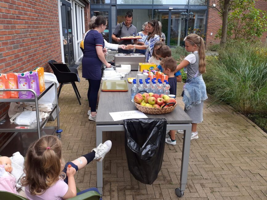 Mensen staan buiten aan tafels met eten en drinken. Kinderen en volwassenen praten en pakken snacks en drankjes.