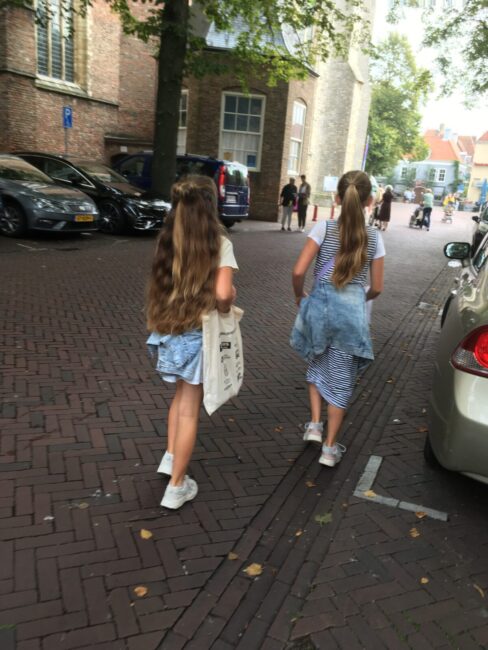 Twee meisjes met lang haar lopen op een geplaveide straat, omringd door geparkeerde auto's en historische gebouwen.