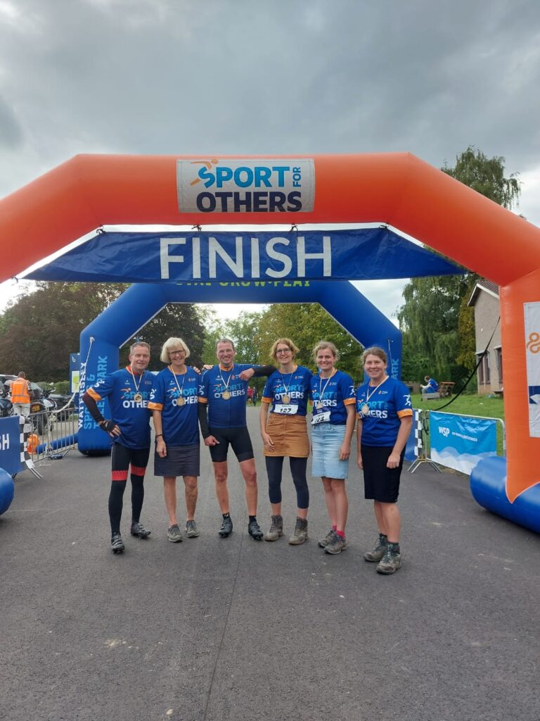 Groep mensen in sportkleding poseert onder een finishboog met de tekst "Sport for Others" op een bewolkte dag.