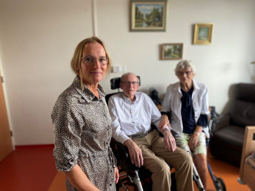 Een vrouw staat naast een oudere man in een rolstoel en een oudere vrouw. Ze zijn in een kamer met schilderijen aan de muur.