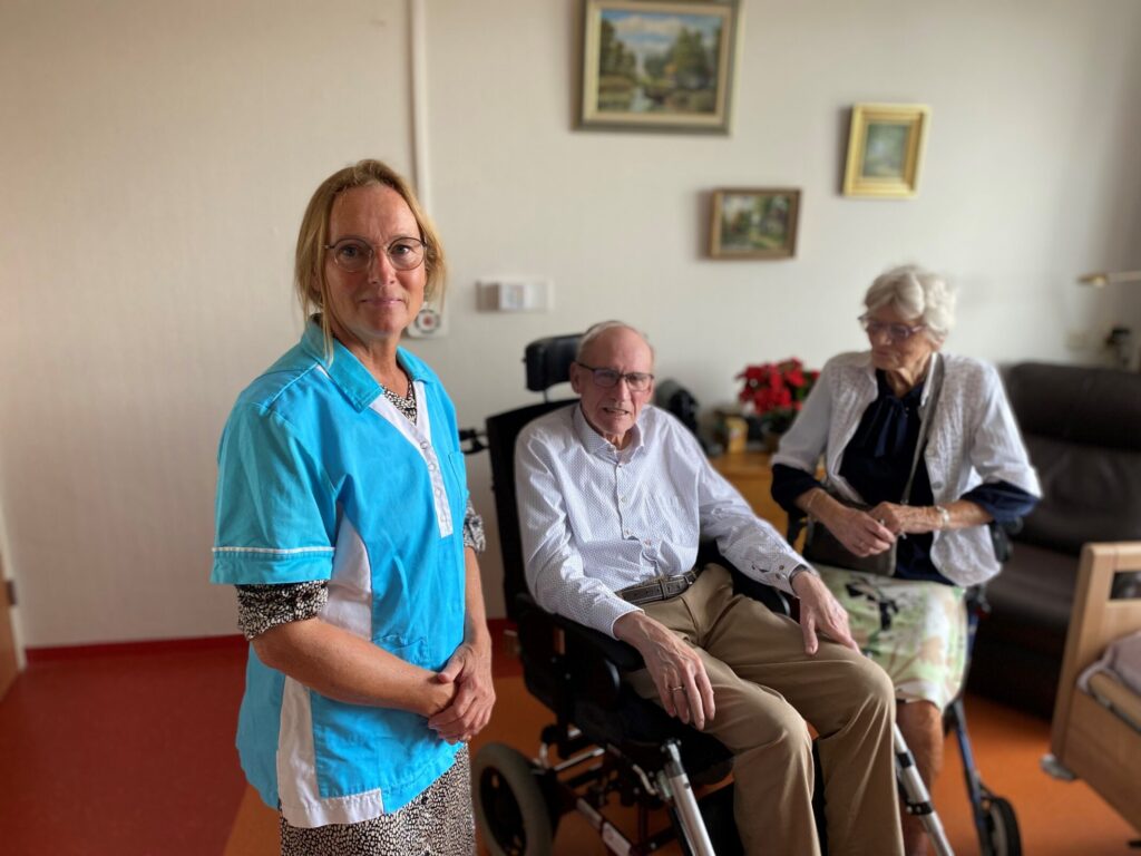 Zorgmedewerker in blauwe uniform staat naast een oudere man in een rolstoel en een oudere vrouw in een woonkamer.