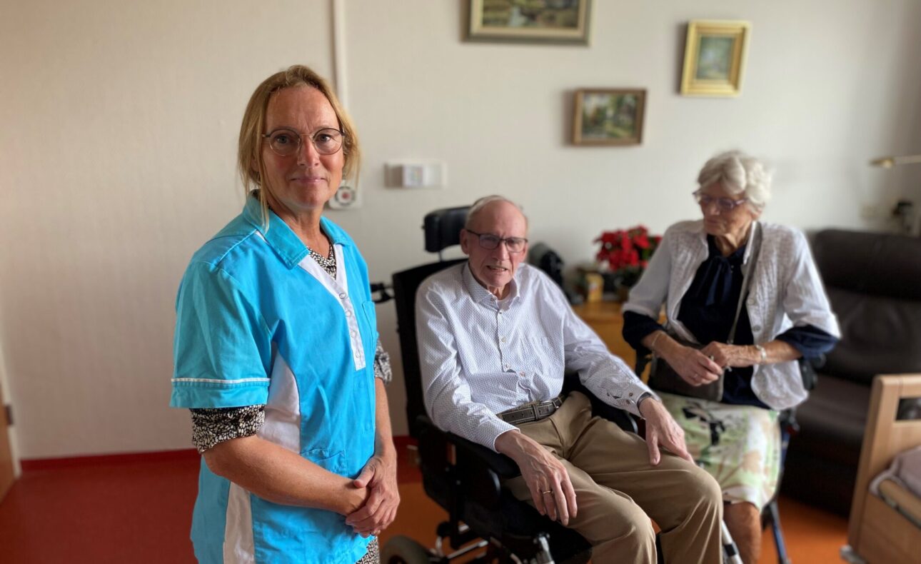 Zorgmedewerker in blauwe uniform staat naast een oudere man in een rolstoel en een oudere vrouw in een woonkamer.