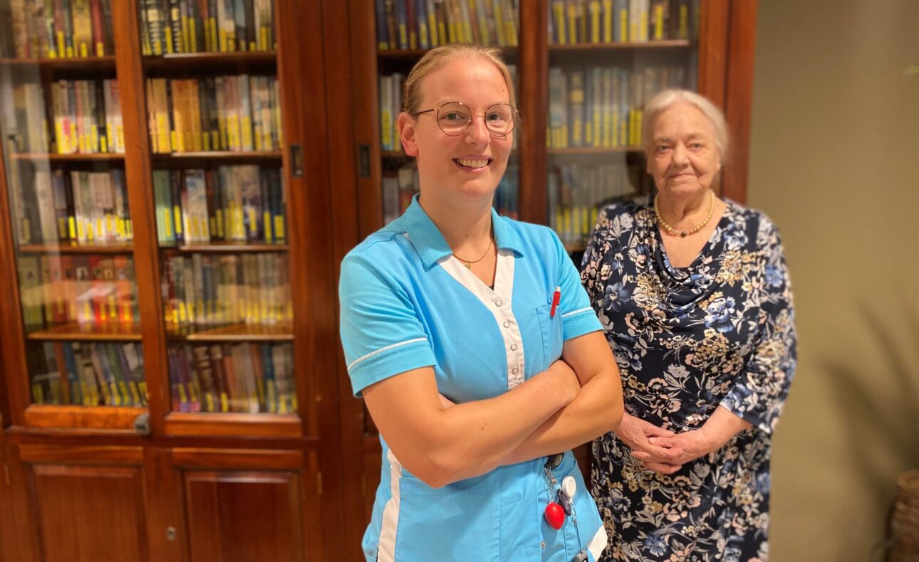 Twee vrouwen staan voor een boekenkast. De vrouw links draagt een blauw uniform, de vrouw rechts een bloemenjurk.