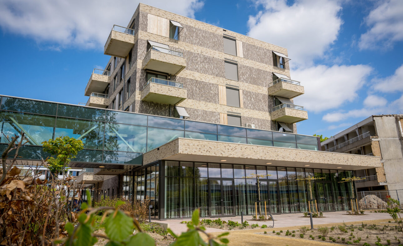 Modern gebouw met glazen gevel en meerdere verdiepingen, omgeven door groen en een heldere blauwe lucht met enkele wolken.