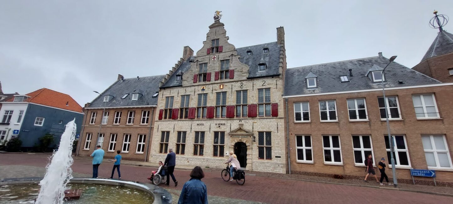 Historisch gebouw met trapgevel en rode luiken op een plein, omringd door mensen en een fontein op de voorgrond.
