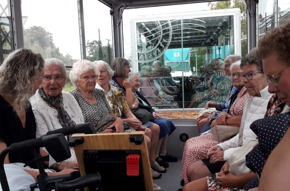 Groep ouderen zit in een overdekte boot met glazen wanden, varend door een groene omgeving. Ze lijken ontspannen en vrolijk.
