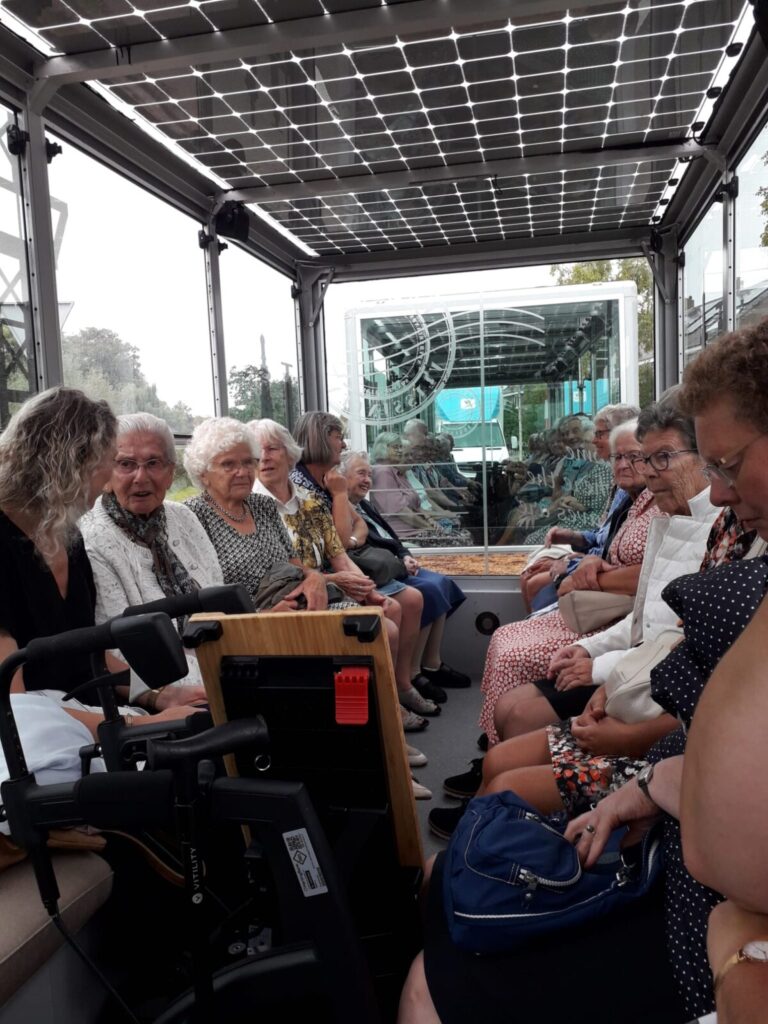 Groep ouderen zit in een overdekte boot met glazen wanden, varend door een groene omgeving. Ze lijken ontspannen en vrolijk.