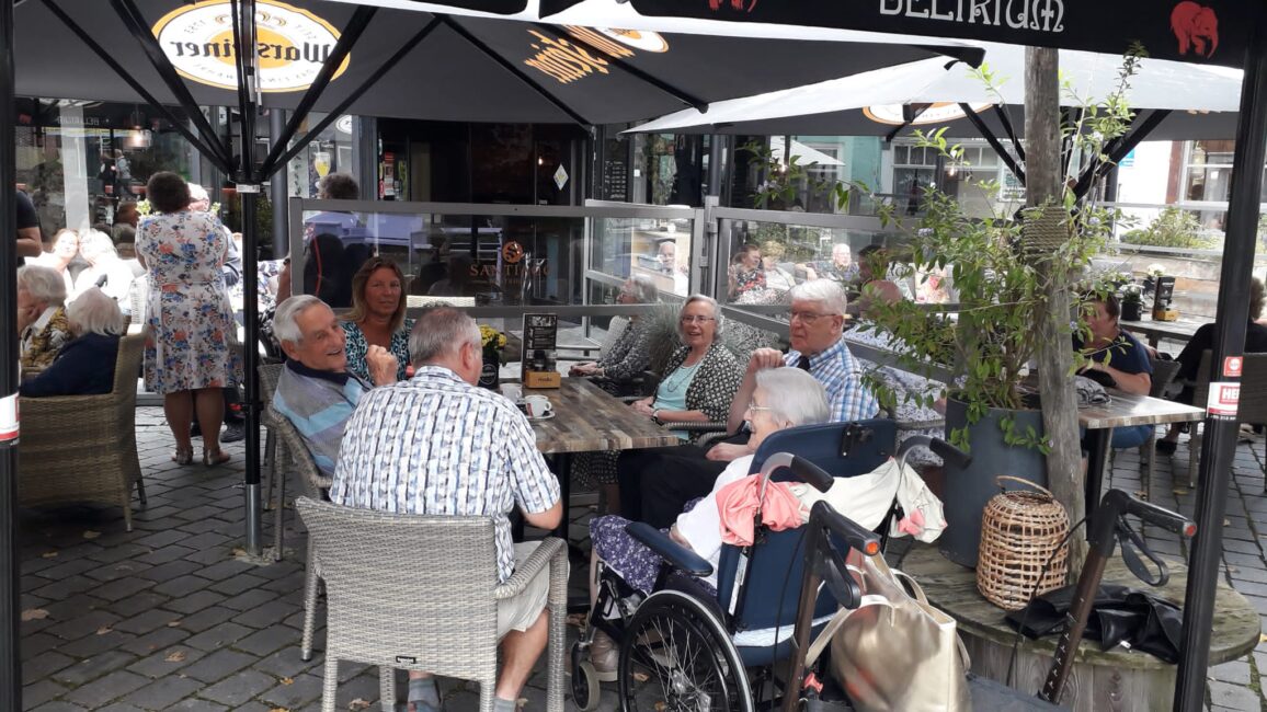 Groep oudere mensen zit gezellig aan een tafel op een terras onder parasols, genietend van een zonnige dag in de buitenlucht.