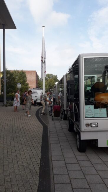 Rij elektrische voertuigen geparkeerd langs een stoep met mensen die in de buurt lopen, onder een bewolkte hemel.