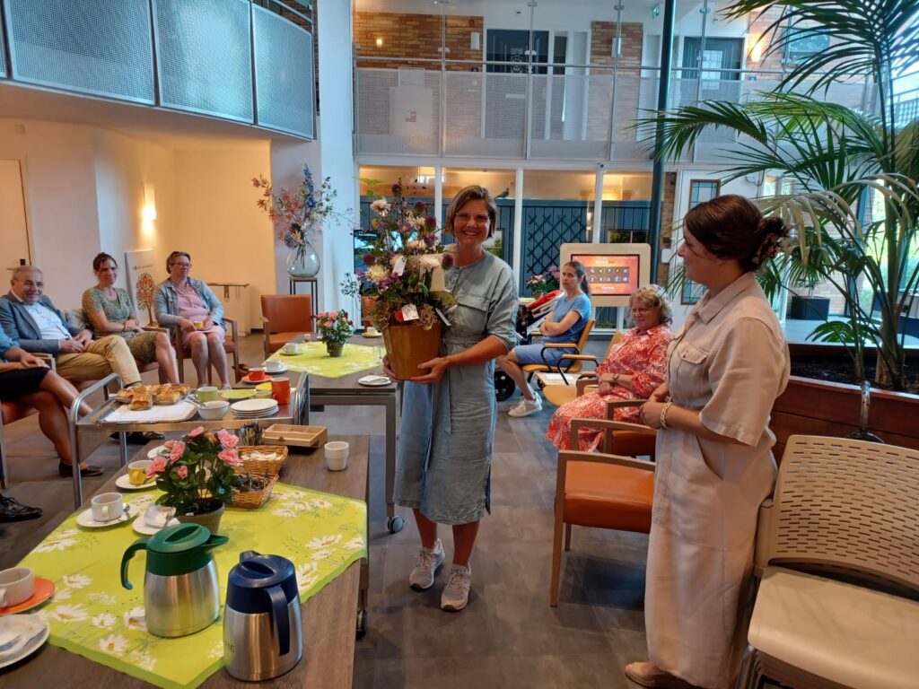 Groep mensen in een lichte ruimte, zittend rond een tafel met bloemen en hapjes. Twee vrouwen staan in het midden met een cadeau.