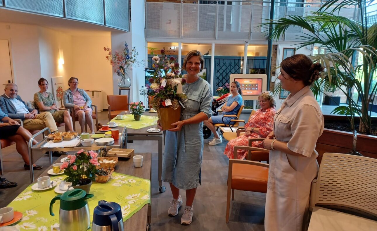 Groep mensen in een lichte ruimte, zittend rond een tafel met bloemen en hapjes. Twee vrouwen staan in het midden met een cadeau.
