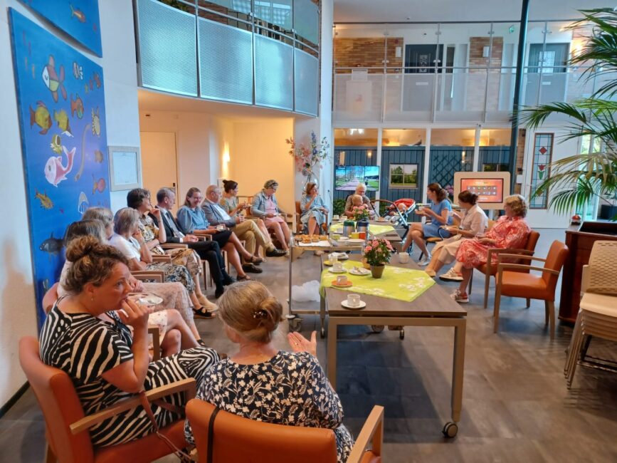 Groep mensen zit in een kring in een lichte ruimte met kunst aan de muur. Er staan tafels met bloemen en drankjes in het midden.