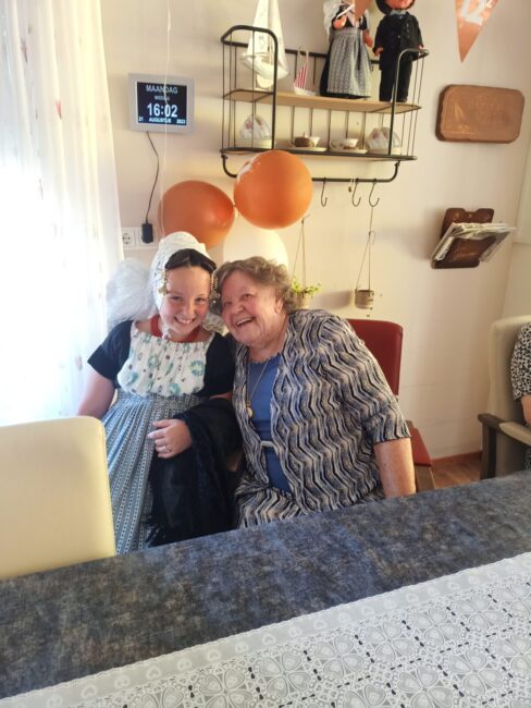 Twee vrouwen zitten glimlachend aan een tafel in een gezellig ingerichte kamer met oranje ballonnen en decoraties aan de muur.