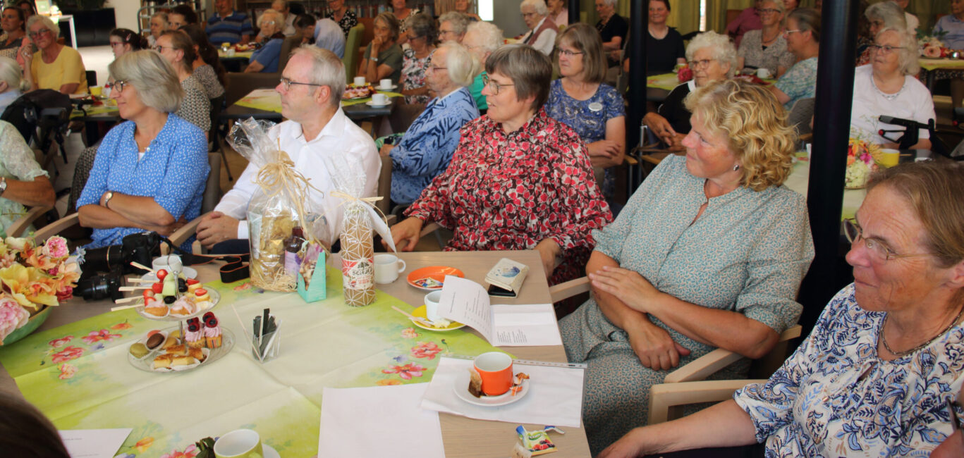 Groep oudere mensen zit aan tafels met hapjes en drankjes tijdens een bijeenkomst in een zaal, aandachtig luisterend.
