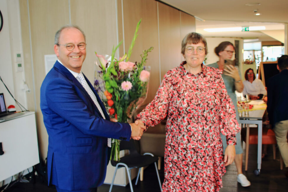 Twee mensen schudden elkaar de hand in een kamer. De man links draagt een blauw pak en houdt een boeket bloemen vast.