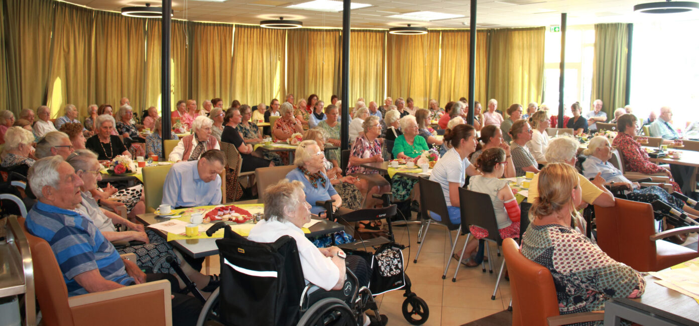 Een grote groep ouderen zit in een zaal met tafels en stoelen, luisterend naar een presentatie of evenement.