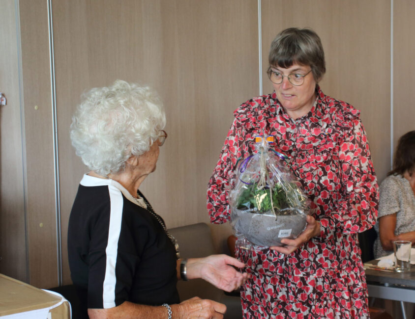 Twee vrouwen in gesprek; de een geeft de ander een cadeau met bloemen in een mand, in een kamer met houten panelen.