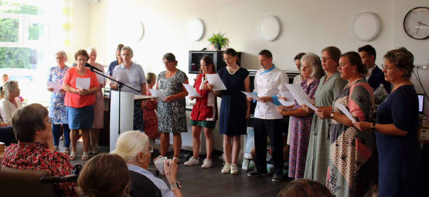 Groep mensen zingt voor een publiek in een lichte kamer. Een spreker staat bij een microfoon. Er hangt kunst aan de muur.
