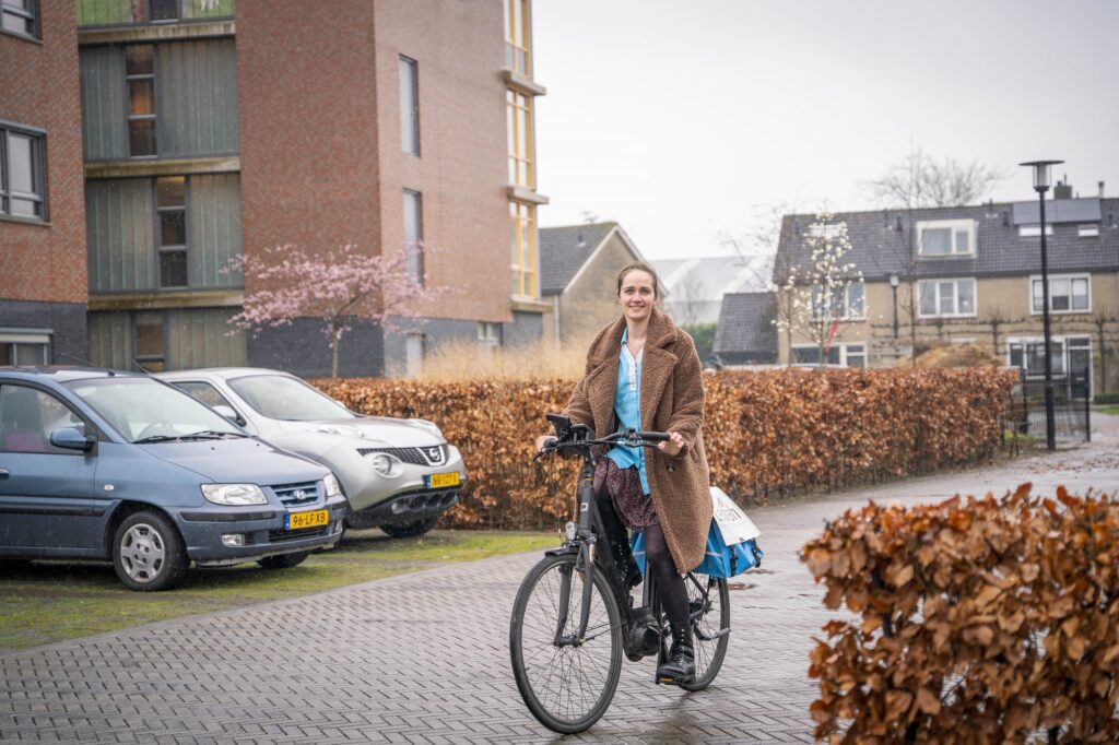 Vrouw fietst op een elektrische fiets op een bestraat pad langs geparkeerde auto's en gebouwen op een bewolkte dag.