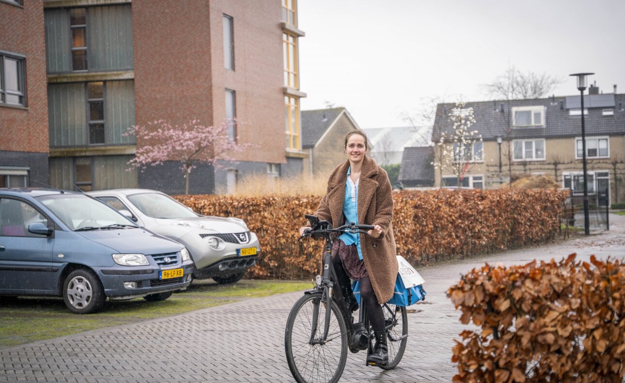 Vrouw fietst op een elektrische fiets op een bestraat pad langs geparkeerde auto's en gebouwen op een bewolkte dag.