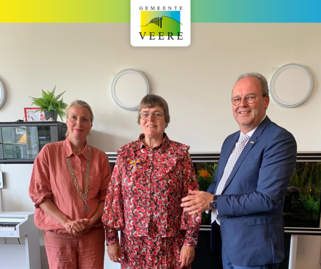 Drie mensen poseren voor een foto in een kamer met een aquarium. De vrouw in het midden draagt een koninklijke onderscheiding.