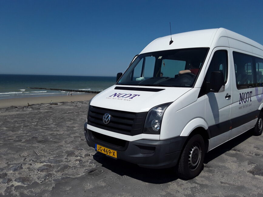 Witte Volkswagen-bus met "Noot" op de zijkant geparkeerd op een strandweg met uitzicht op de zee en een blauwe lucht.
