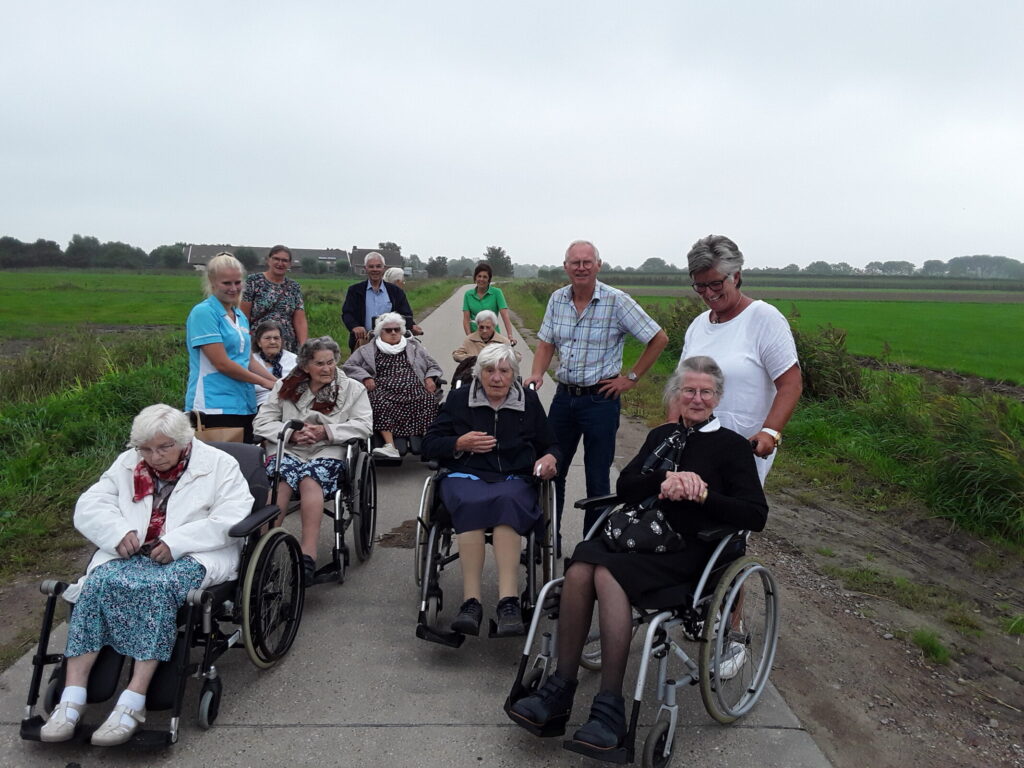 Groep ouderen en begeleiders op een landweg, sommigen in rolstoelen. Op de achtergrond is een groen veld zichtbaar.