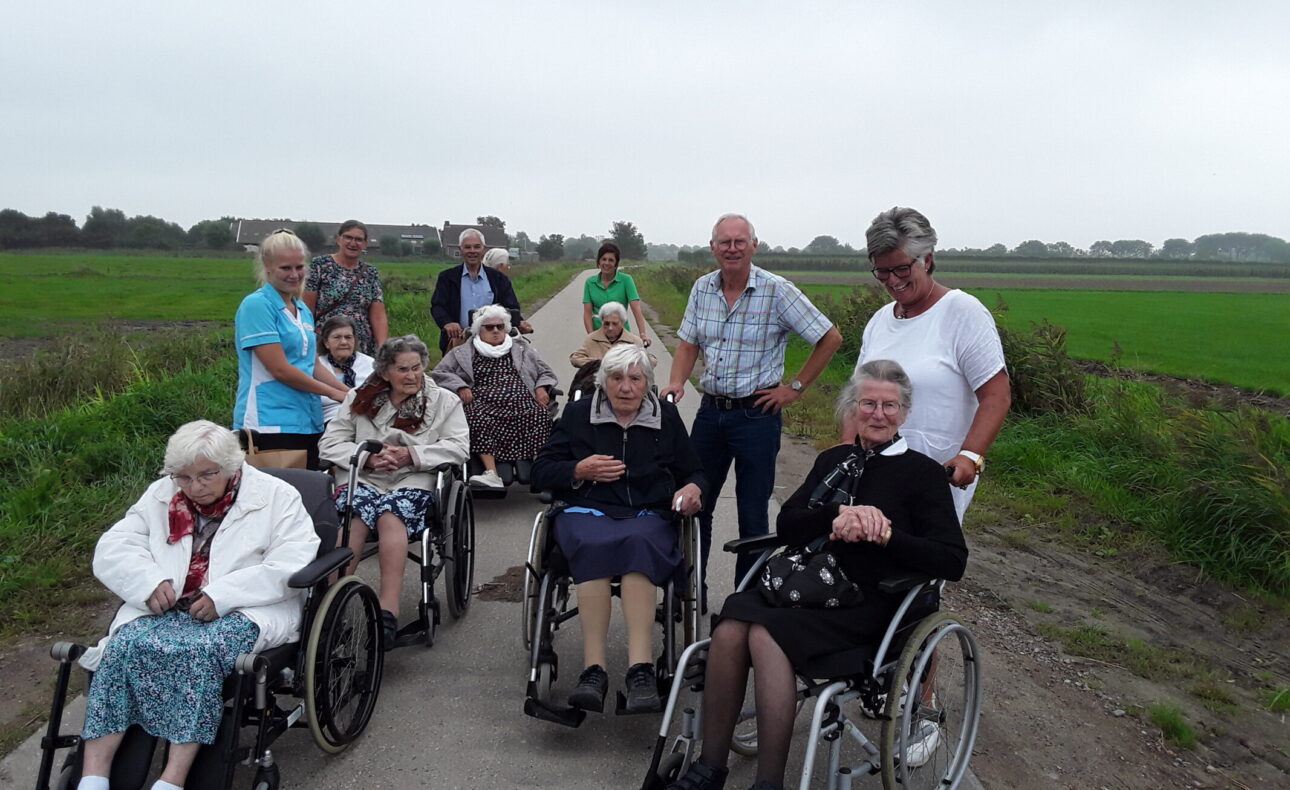 Groep ouderen en begeleiders op een landweg, sommigen in rolstoelen. Op de achtergrond is een groen veld zichtbaar.