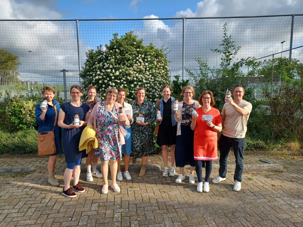 Groep mensen poseert buiten voor een groene struik, met een hek op de achtergrond. Ze houden bekers vast en lachen.