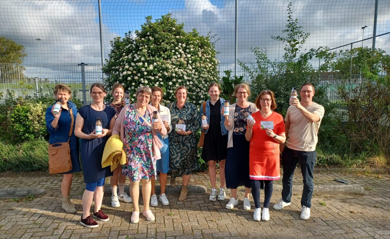 Groep mensen poseert buiten voor een groene struik, met een hek op de achtergrond. Ze houden bekers vast en lachen.