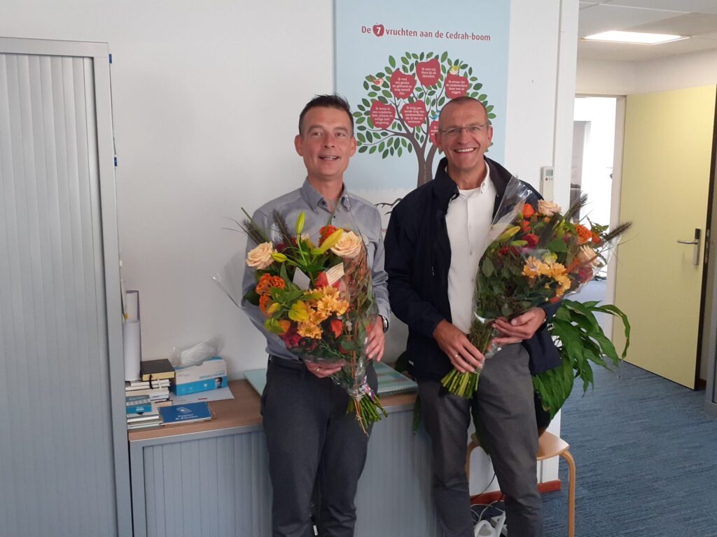 Twee mannen staan binnen en houden boeketten vast. Op de achtergrond is een schilderij van een boom met rode bloemen te zien.