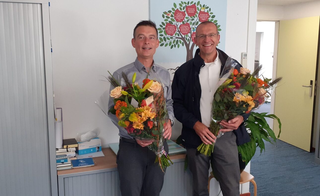 Twee mannen staan binnen en houden boeketten vast. Op de achtergrond is een schilderij van een boom met rode bloemen te zien.