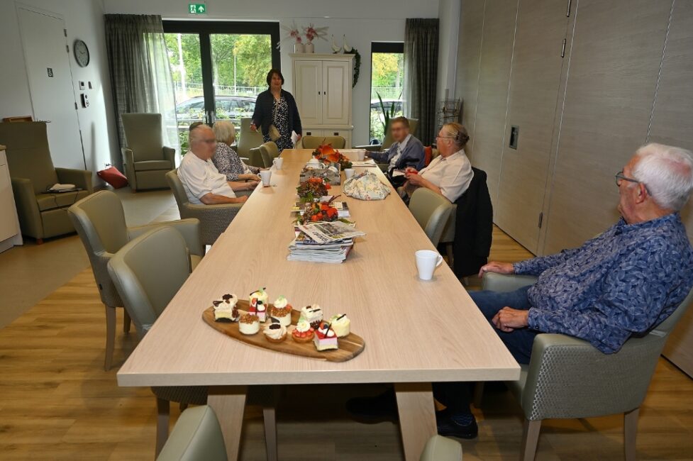 Een groep ouderen zit aan een lange tafel in een gemeenschappelijke ruimte, met gebakjes en koffie op tafel.