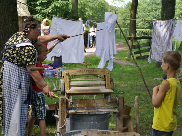 Een vrouw en kinderen doen de was met een ouderwetse wasmachine in een groene buitenomgeving, met wasgoed aan de lijn.