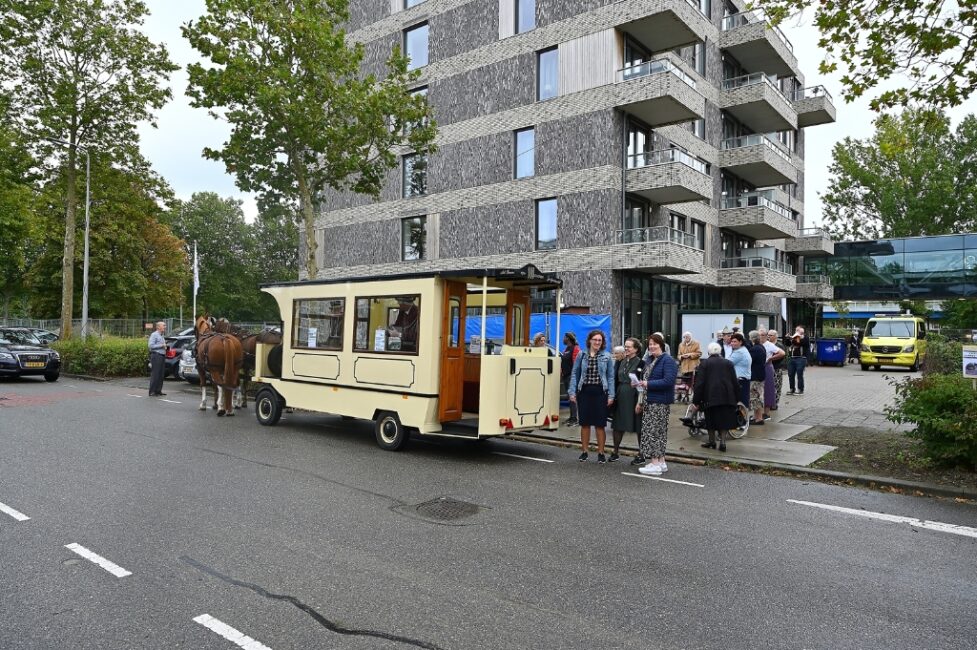 Een paardentram staat geparkeerd voor een modern gebouw. Mensen staan op de stoep en kijken naar de tram en het paard.