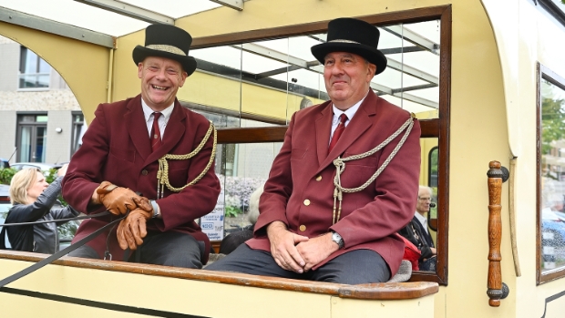 Twee mannen in bordeauxrode uniformen en hoge hoeden zitten lachend op een koets. Ze dragen handschoenen en hebben touwen vast.