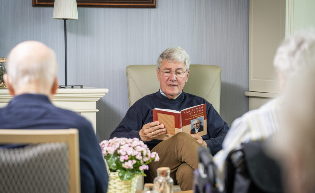 Een oudere man leest voor uit een boek aan een groep mensen in een gezellige kamer met bloemen en een lamp op de achtergrond.