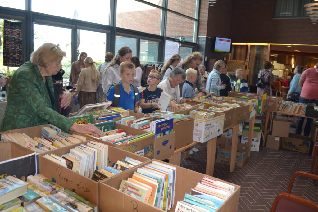 Mensen bladeren door boeken op een boekenmarkt in een ruime, lichte hal. Tafels zijn gevuld met boeken in dozen.