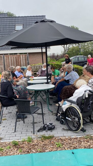 Groep ouderen zit buiten rond een tafel onder een parasol. Sommigen zitten in rolstoelen. Op de achtergrond is een huis te zien.