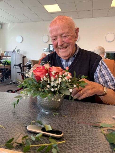 Oudere man met een glimlach schikt een boeket bloemen in een vaas op tafel in een lichte kamer met een rolstoel op de achtergrond.