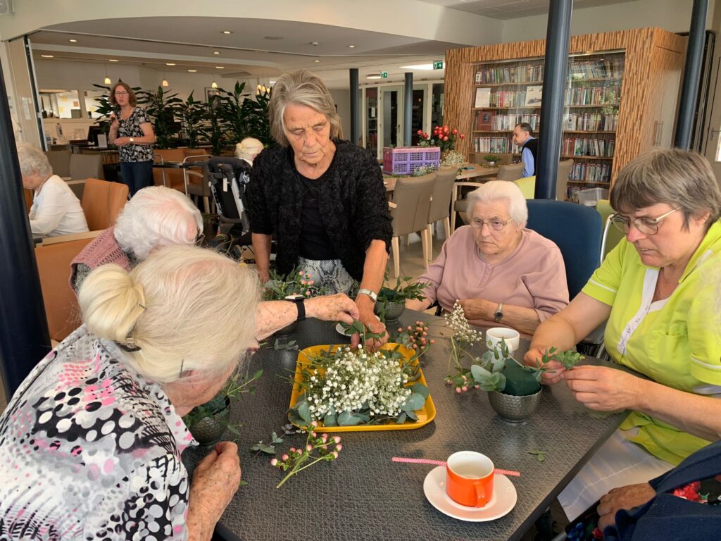 Een groep ouderen en een begeleider maken bloemstukken aan een tafel in een gemeenschappelijke ruimte met boekenplanken.
