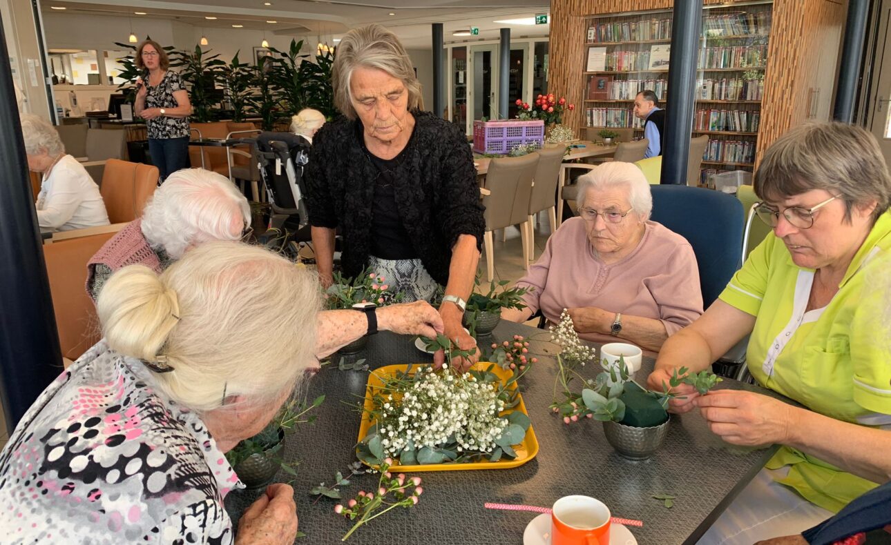 Een groep ouderen en een begeleider maken bloemstukken aan een tafel in een gemeenschappelijke ruimte met boekenplanken.