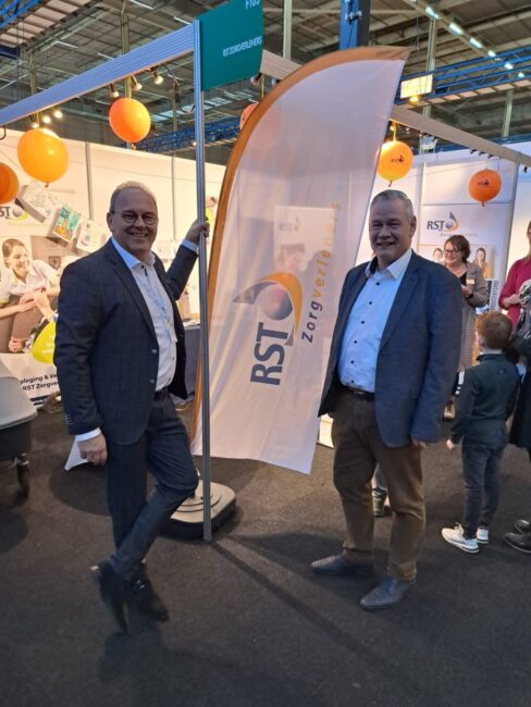 Twee mannen in pakken staan naast een vlag met het logo van RST Zorg. Op de achtergrond zijn ballonnen en een beursstand.