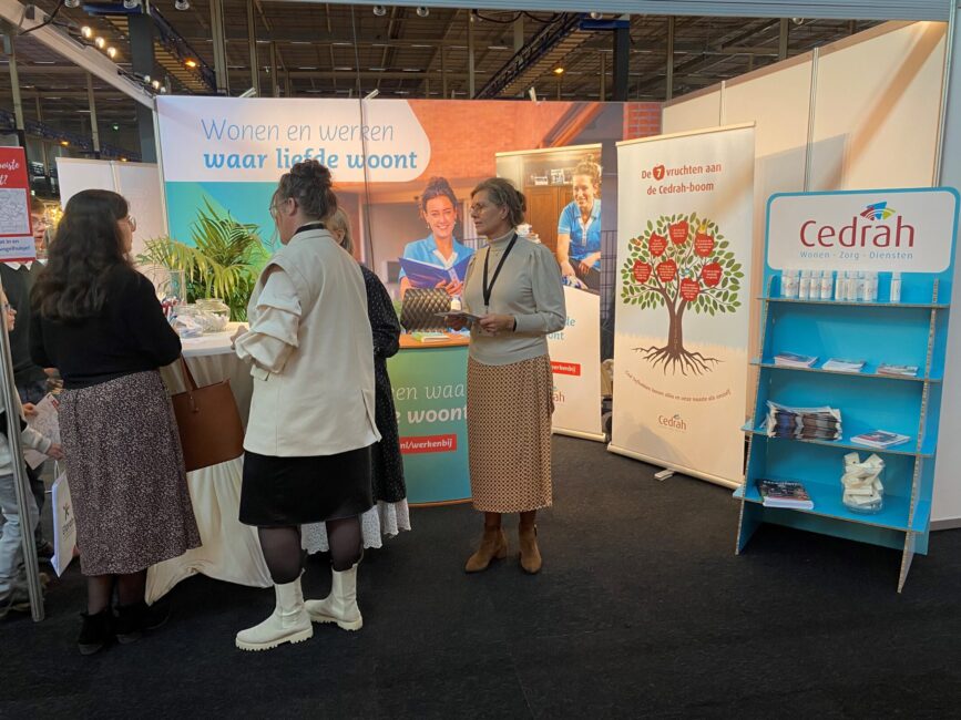 Drie vrouwen staan bij een informatiestand van Cedrah op een beurs. Op de achtergrond zijn banners en folders te zien.