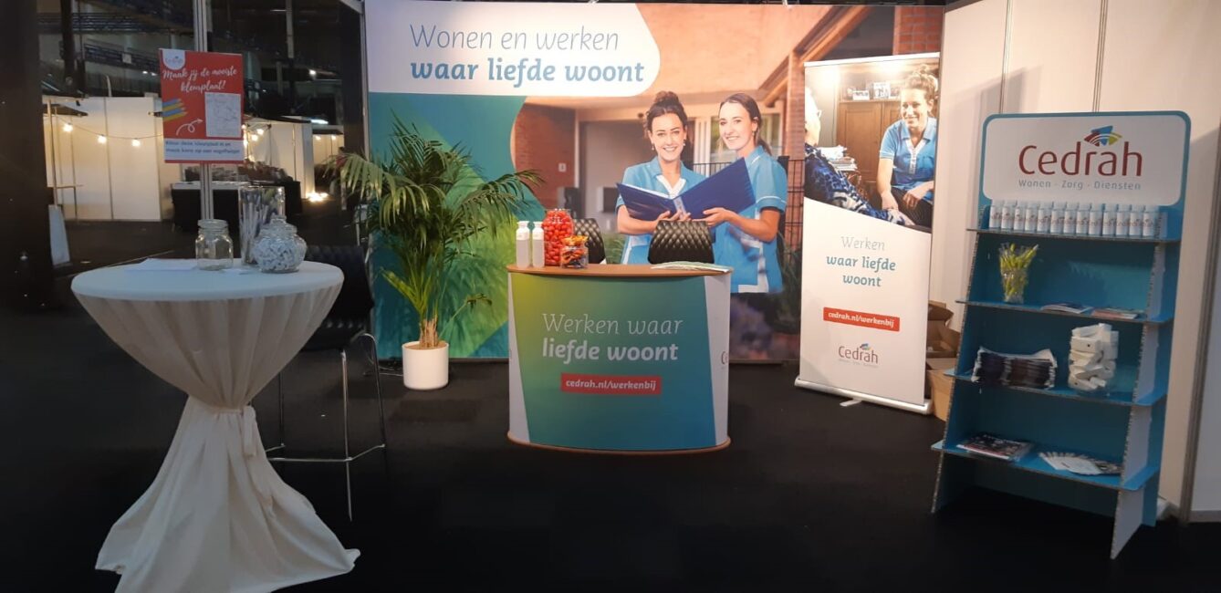 Beursstand van Cedrah met promotiemateriaal, brochures en banners. Een tafel met decoratie staat links van de stand.