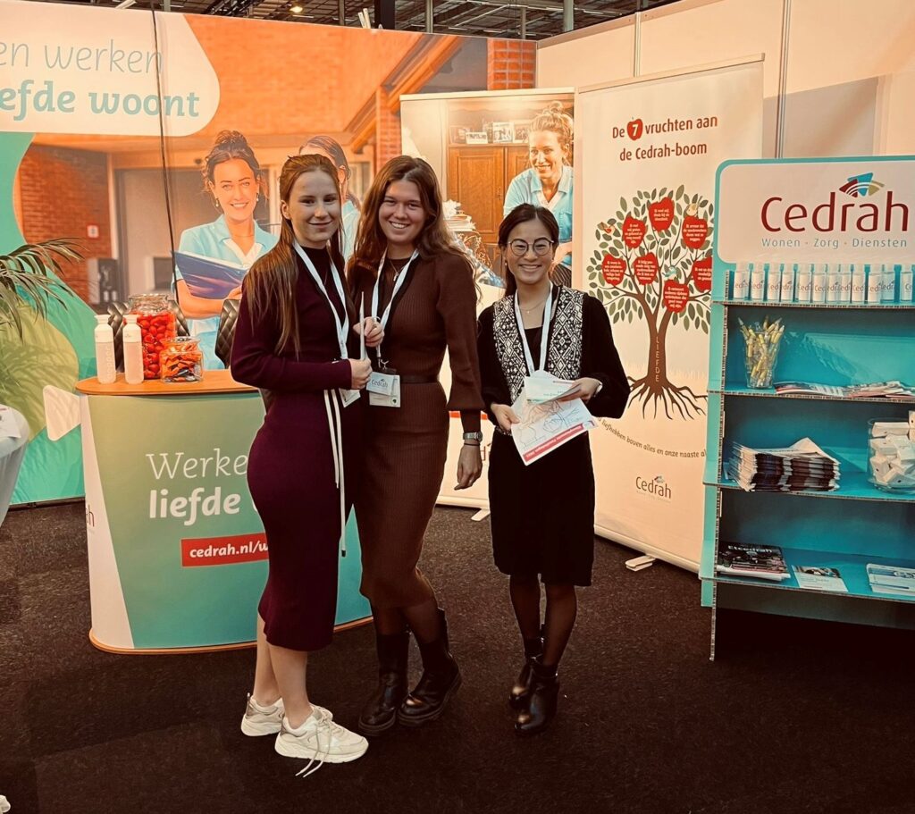 Drie vrouwen staan glimlachend voor een Cedrah-stand op een beurs, met promotiemateriaal en banners op de achtergrond.