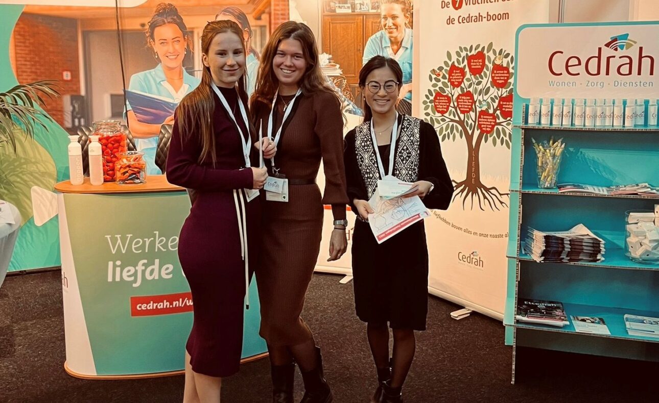 Drie vrouwen staan glimlachend voor een Cedrah-stand op een beurs, met promotiemateriaal en banners op de achtergrond.