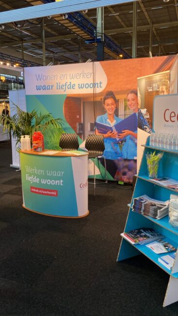 Beursstand met promotiemateriaal en brochures van Cedrah, met de slogan "Werken waar liefde woont" op de achtergrond.